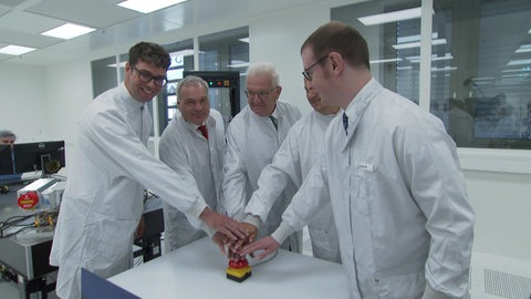 Ministerpräsident Winfried Kretschmann (Grüne) trifft sich mit Mitarbeitenden der Backnanger Firma Tesat in einem Unternehmensgebäude und trägt dabei wie sie einen weißen Laboranzug.