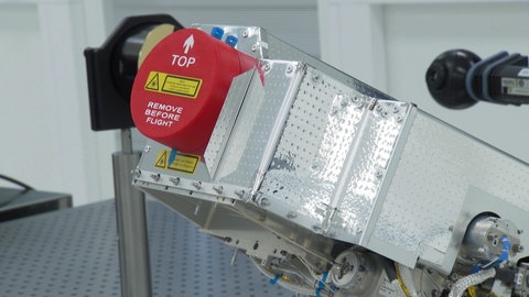 Ministerpräsident Winfried Kretschmann (Grüne) trifft sich mit Mitarbeitenden der Backnanger Firma Tesat in einem Unternehmensgebäude und trägt dabei wie sie einen weißen Laboranzug.