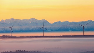 Drei Windräder ragen am Morgen kurz nach Sonnenaufgang aus der Nebeldecke, während im Hintergrund durch den Föhn die über einhundert Kilometer entfernten Alpen zu sehen sind.