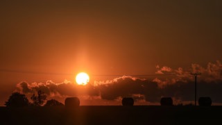Strohballen liegen auf einem Feld, während die Sonne am Horizont untergeht.