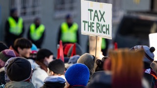 Auf einem Schild steht bei einer Demonstration steht "tax the rich". In einem offenen Brief fordern Superreiche für höhere Steuern für sich.