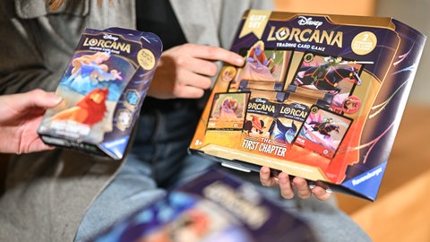 Zwei Mitarbeiterinnen des Spieleherstellers Ravensburger AG halten das neue Sammelkartenspiel Disney Lorcana Trading Card Game, The First Chapter in den Händen. Das Sammelkartenspiel wird von der Ravensburger AG hergestellt und beschert dem Unternehmen Umsätze in Millionen-Höhe.
