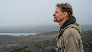 Lars steht auf einem Hügel an der Küste auf der schottischen Insel Colonsay und schaut in die Weite.