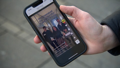 Eine Hand hält ein Smartphone, darauf ist ein TikTok-Video des BW-Finanzministeriums mit tanzenden Personen und der Text "Das Finanzamt auf dem Weg um deine Nachzahlung zu holen" zu sehen.