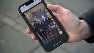 Eine Hand hält ein Smartphone, darauf ist ein TikTok-Video des BW-Finanzministeriums mit tanzenden Personen und der Text "Das Finanzamt auf dem Weg um deine Nachzahlung zu holen" zu sehen.