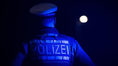 Ein Polizist steht mit dem Rücken zur Kamera im Dunkeln. Auf seiner Jacke ist das Wort "Polizei" zu lesen. (Symbolbild)