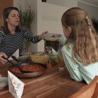 Eine Frau in gestriftem Oberteil reicht einem Mädchen im gepunkteten Kleid einen Teller mit Nudeln und Soße. Vera Maurer sagt, sie habe früher nicht geglaubt, dass sie einmal ein Leben als Hausfrau und Mutter führen würde.
