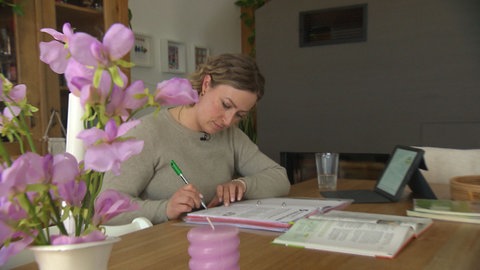 Eine blonde Frau sitzt an einem Esstisch und schreibt in ein Ringbuch, vor ihr liegt ein Tablet, im Vordergrund steht eine Pflanze: Für Lehrerin Silvia Wolter gibt es auch zu Hause immer etwas für die Schule zu tun.