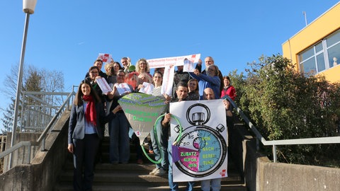 Menschen stehen mit Transparenten auf einer Treppe. Eine Aktion der GEW im Main-Tauber-Kreis: Die Bildungsgewerkschaft fordert eine Arbeitszeiterfassung für Lehrkräfte.