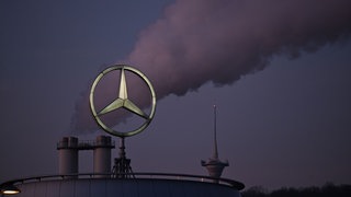 Der Mercedes Stern, das Logo des Fahrzeugherstellers Mercedes-Benz steht auf einem Gebäude einer Niederlassung, dahinter steigt eine Wolke aus einem Schornstein auf. Laut dem Handelsblatt soll der Autobauer hohe sechsstellige Summen an Abfindungen bezahlen.
