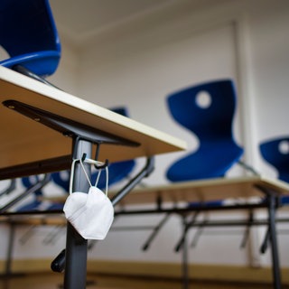 Symbolbild | Mundschutzmaske hängt an einem Schultisch in einem Klassenzimmer mit auf den Tischen aufgestellten Stühlen.
