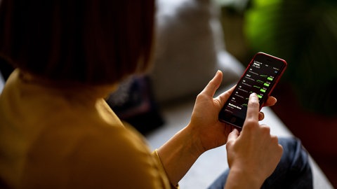 Eine junge Frau beobachtet mit ihrem Smartphone die Aktienkurse in einer Trading-App. Durch den niederschwelligen Einstieg investieren immer mehr junge Menschen an der Börse.
