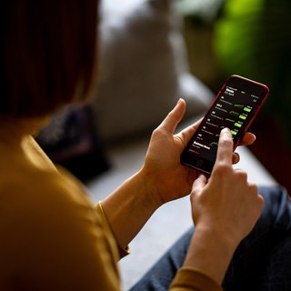 Eine junge Frau beobachtet mit ihrem Smartphone die Aktienkurse in einer Trading-App. Durch den niederschwelligen Einstieg investieren immer mehr junge Menschen an der Börse.