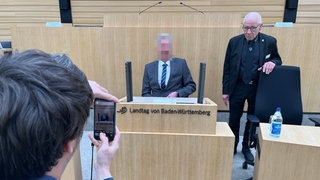 Der Untersuchungsausschuss des baden-württembergischen Landtages zur Polizei-Affäre geht weiter. Am Montag gab der ehemalige Inspekteur eine Erklärung ab. Das Bild zeigt ihn verpixelt in der Mitte, der Mann neben ihm gehört zum Sicherheitspersonal im Landtag. 