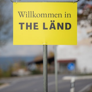 An einem Ortsschild hängt ein Zusatzschild mit der Aufschrift "Willkommen in THE LÄND2.