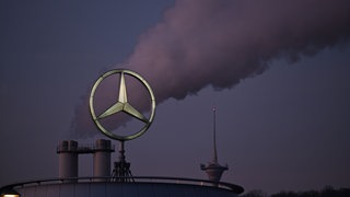 (Symbolbild) Der Mercedes Stern, das Logo des Fahrzeugherstellers Mercedes-Benz steht auf einem Gebäude einer Niederlassung, dahinter steigt eine Wolke aus einem Schornstein auf.