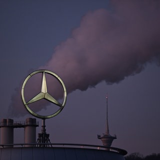 (Symbolbild) Der Mercedes Stern, das Logo des Fahrzeugherstellers Mercedes-Benz steht auf einem Gebäude einer Niederlassung, dahinter steigt eine Wolke aus einem Schornstein auf.
