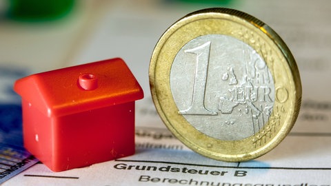 Eine Euro-Geldmünze sowie ein Spielzeughaus stehen auf einem Abgabenbescheid für die Entrichtung der Grundsteuer.