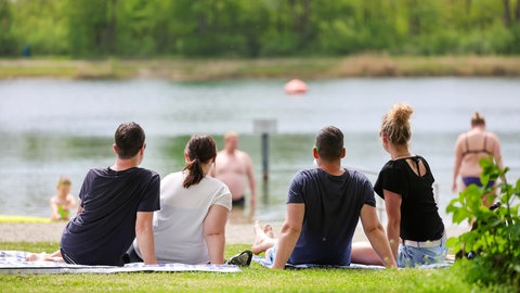 Menschen sitzen im Sonnenschein am Ufer des Schwarzachtalsees bei Ertingen (Kreis Biberach), im Hintergrund sind Badegäste im Wasser.