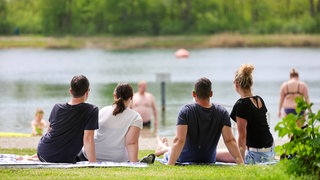 Menschen sitzen im Sonnenschein am Ufer des Schwarzachtalsees bei Ertingen (Kreis Biberach), im Hintergrund sind Badegäste im Wasser.