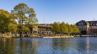 Menschen vor dem Landtag in Stuttgart, dem Zentrum der BW-Politik, im Vordergrund der Eckensee.