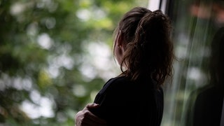 Eine junge Frau steht in einer Wohnung und schaut durch ein Fenster nach draußen.