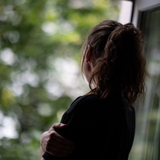 Eine junge Frau steht in einer Wohnung und schaut durch ein Fenster nach draußen.