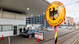 Ein Grenzschild steht am Grenzübergang in Weil am Rhein - Friedlingen während im Hintergrund zwei Beamte der Bundespolizei zu sehen sind. 