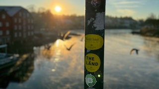 An einem Pfahl auf einer Brücke kleben Aufkleber mit der Aufschrift "Nett hier" und "The Länd".