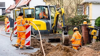 Arbeiter sind mit Bagger und Spaten an einer Straße mit der Erdverlegung von Glasfaserkabeln beschäftigt.