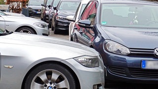Symbolbild: Mehrere geparkte Autos blockieren einen Bürgersteig, den man kaum noch erkennen kann. Städte und Gemeinden kämpfen verstärkt mit Falschparkern an Badeseen in Baden-Württemberg.