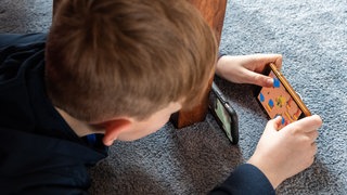 Ein Junge spielt ein Videospiel auf seinem Smartphone und überträgt es mit einem zweiten Handy per Facetime zu seinem Freund. 