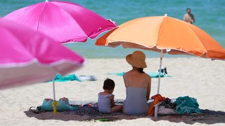Eine Frau und ein Kind sitzen am Strand unter einem Sonnenschirm.