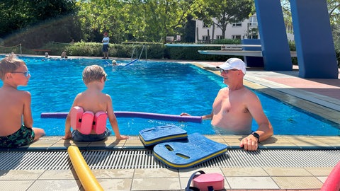Zwei Jungen sitzen am Beckenrand eines Schwimmbeckens. Ein Schwimmeister ist neben ihnen im Wasser, er hat Schwimmbretter und -nudeln neben sich.