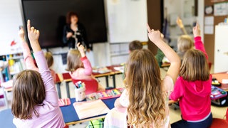 Schüler einer gemischten ersten und zweiten Klasse sitzen während des Unterrichts auf ihren Plätzen und heben die Hände. Mit Blick auf 1.440 unwissentlich unbesetzte Lehrerstellen in Baden-Württemberg gibt es noch viele offenen Fragen.