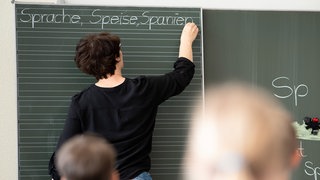 Symbolbild: Eine Lehrerin schreibt in einer Schule Worte an eine Tafel.