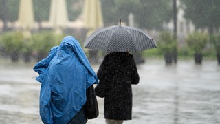 Ein Mensch schützt sich mit einer Jacke vor dem Regen, eine andere Person hat einen Schirm.