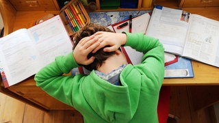 Ein Junge hält sich den Kopf, während er Hausaufgaben macht. Viele Kinder leiden unter Kopfschmerzen. (Symbolbild)