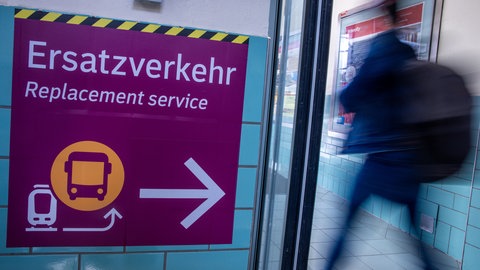 Ein lila Schild weist auf den Ersatzverkehr mit Bussen hin. Ein Fahrgast rennt vorüber.
