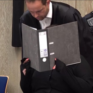 Eine Angeklagte hält sich im Gerichtsaal einen Ordner vors Gesicht