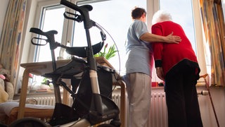 Eine Pflegerin und eine Seniorin stehen am Fenster, im Vordergrund ist ein Rollator zu sehen. Derzeit wird diskutiert, ob der Pflegegrad 1 abgeschafft werden soll, um Geld zu sparen.