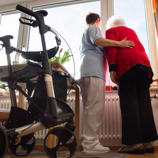 Eine Pflegerin und eine Seniorin stehen am Fenster, im Vordergrund ist ein Rollator zu sehen. Derzeit wird diskutiert, ob der Pflegegrad 1 abgeschafft werden soll, um Geld zu sparen.