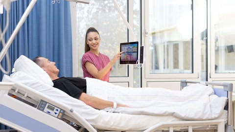 Auf der Inspire Living Lab Station steht eine Krankenpflegerin neben dem Bett eines Patienten und richtet einen Tablet-PC auf ihn aus.