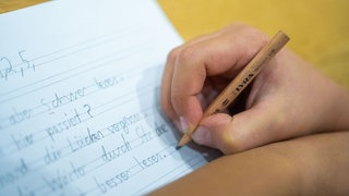 Symbolbild: Ein Grundschüler sitzt an einem Tisch und schreibt mit einem Bleistift Wörter in sein Heft. Beim Kompass 4 Leistungstest werden Grundschülerinnen und Grundschüler neben Mathe auch in Deutsch geprüft. 