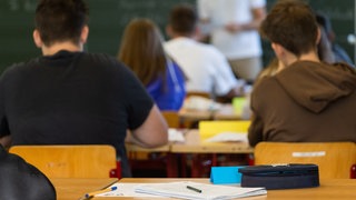 Schüler sitzen in einem Klassenzimmer, an der Tafel steht ein Lehrer.