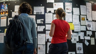 Zwei Studentinnen betrachten am die Wohnungsanzeigen am Schwarzen Brett. Ein WG-Zimmer zu finden, ist in vielen Städten in Baden-Württemberg schwer.