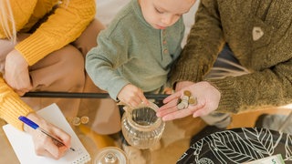Ein Junge zählt Münzen, rechts und links von ihm sitzen seine Eltern (von oben betrachtet). Wie können Familien effektiv Geld anlegen?