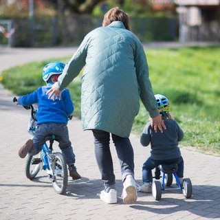 Symbolbild | Eine Mutter schiebt ihre kleinen Kinder an, die mit Fahrrad und Tretroller unterwegs sind. 