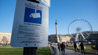 Ein Schild zeigt den Hinweis auf zeitlich beschränkte Videoüberwachung. Im Hintergrund ist der Stuttgarter Schlossplatz zu sehen. 