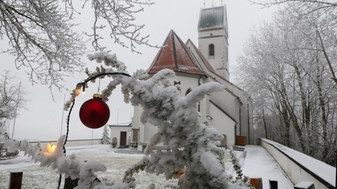 Eine rote Christbaum-Kugel hängt an einem eingeschneiten Ast vor der Wallfahrtskirche St. Johannes Baptist in Oberschwaben. Zum ersten Mal nach zehn Jahren gibt es in vielen Regionen Baden-Württembergs weiße Weihnachten.